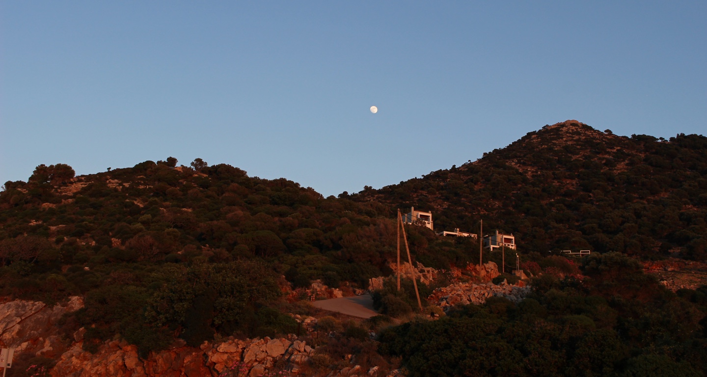 The moon above a quiet hillside, with houses and rocks catching the last warm light.